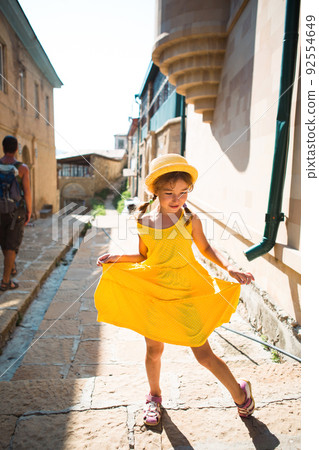 A tourist girl in a yellow hat and sundress dancing along the street of the old town made of stone with a fortress. Sightseeing tour. 92554649
