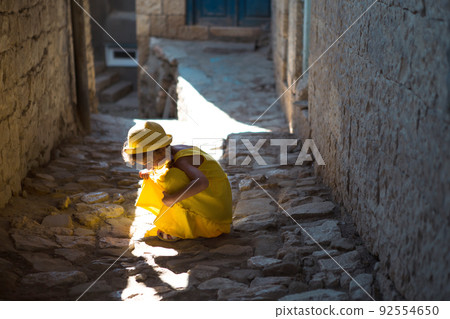 A tourist girl in a yellow hat and sundress walks along the street of the old town made of stone with a fortress. Sightseeing tour. The child got lost, searching for parents 92554650