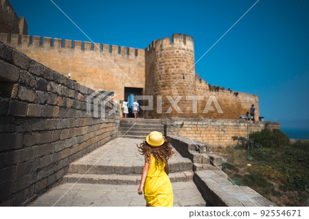 A tourist girl in a yellow hat and sundress walks along the street of the old town made of stone with a fortress. Sightseeing tour. The child got lost, searching for parents 92554671