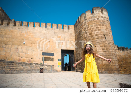 A tourist girl in a yellow hat and sundress walks along the street of the old town made of stone with a fortress. Sightseeing tour. The child got lost, searching for parents 92554672