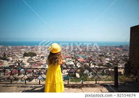 A tourist girl in a yellow hat and sundress looks from a height on the observation deck at the city. Sightseeing tour. The child got lost, searching for parents 92554674
