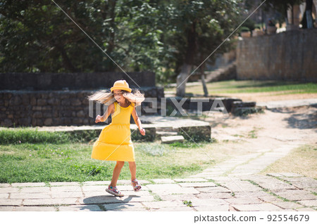A tourist girl in a yellow hat and sundress dancing along the street of the old town made of stone with a fortress. Sightseeing tour. 92554679