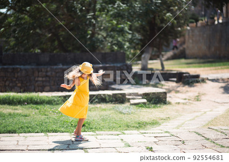 A tourist girl in a yellow hat and sundress dancing along the street of the old town made of stone with a fortress. Sightseeing tour. 92554681