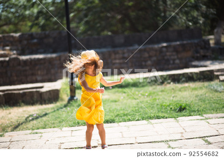 A tourist girl in a yellow hat and sundress dancing along the street of the old town made of stone with a fortress. Sightseeing tour. 92554682