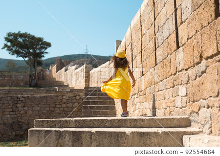 A tourist girl in a yellow hat and sundress walks along the street of the old town made of stone with a fortress. Sightseeing tour. The child got lost, searching for parents 92554684