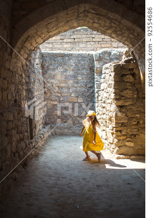 A tourist girl in a yellow hat and sundress dancing along the street of the old town made of stone with a fortress. Sightseeing tour. 92554686