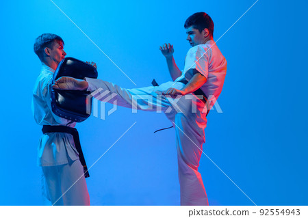 Studio shot of sports training of two karatedo fighters in doboks isolated on blue background in neon. Concept of combat sport, challenges, skills 92554943