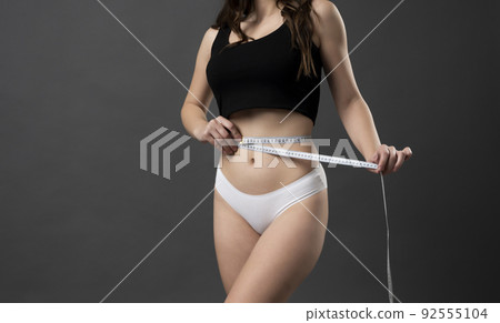 Attractive sexy sporty slim woman body in a black top and white shorts with measuring tape around the waist. Healthy nutrition and weight losing concept. Diet concept. 92555104