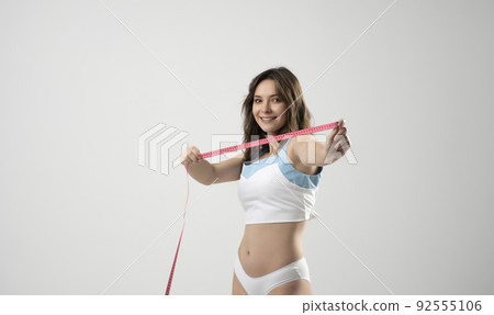 Happy sexy brunette fitness woman with pink measure tape before measuring her waistline on white background. Time for diet and slimming weight loss. Health care and healthy nutrition. 92555106