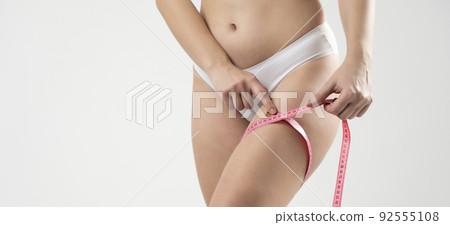 Young slim woman in a white shorts measures her thigh with a pink measuring tape. Healthy food and diet concept. 92555108