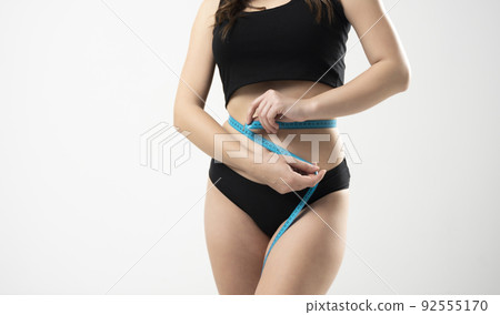 Front view of woman measuring her waist with a blue measuring tape. Losing weight. A girl measures her waist with a centimeter tape. Ideal figure. Slim stomach. Front view of woman measuring her waist with a blue measuring tape. Losing weight. A girl measures her waist with a centimeter tape. Ideal figure. Slim stomach. 92555170
