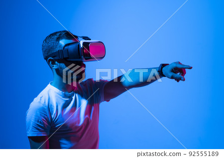 Man in white t-shirt wearing VR goggles using metaverse technology innovation in the virtual world. AR Concept over neon lighting and dark background. Virtual Reality experience. Future technology. Man in white t-shirt wearing VR goggles using metaverse technology innovation in the virtual world. AR Concept over neon lighting and dark background. Virtual Reality experience. Future technology. 92555189