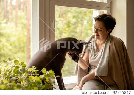 Middle age beautiful woman sitting on windowsill with her dog. 50-year-old woman spending time with her Mexican hairless dog at home. Xoloitzquintle, xolo breed. Dog as best friend, family member. Middle age beautiful woman sitting on windowsill with her dog. 50-year-old woman spending time with her Mexican hairless dog at home. Xoloitzquintle, xolo breed. Dog as best friend, family member. 92555847