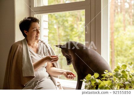 Middle age beautiful woman sitting on windowsill with her dog. 50-year-old woman spending time with her Mexican hairless dog at home. Xoloitzquintle, xolo breed. Dog as best friend, family member. 92555850