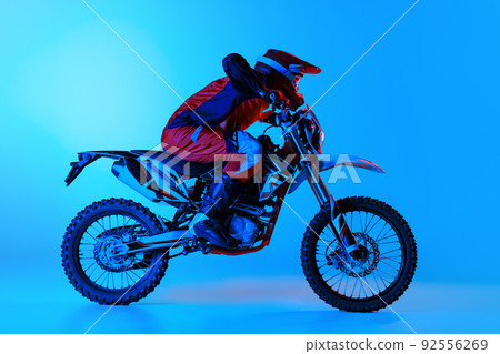 Portrait of young man, biker in full equipment riding motorbike isolated over blue studio background in neon light. Motocross 92556269