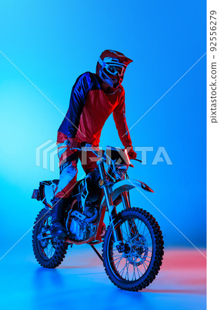 Portrait of young man, biker in full protective equipment standing on motorbike isolated over blue studio background in neon light Portrait of young man, biker in full protective equipment standing on motorbike isolated over blue studio background in neon light 92556279
