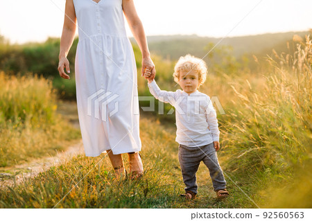 Beautiful woman in light white dress leads her son by the hand along the path of a picturesque field Beautiful woman in light white dress leads her son by the hand along the path of a picturesque field 92560563