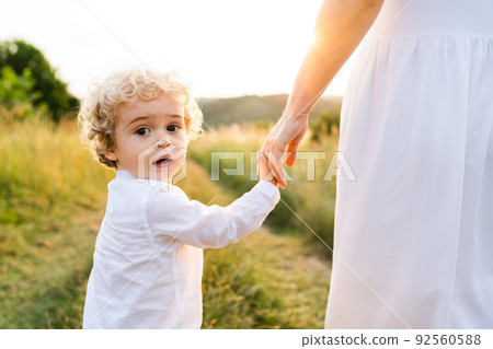 Cute boy with blond curly hair walks with his mother along a picturesque field at sunset and turns back looking at the camera Cute boy with blond curly hair walks with his mother along a picturesque field at sunset and turns back looking at the camera 92560588