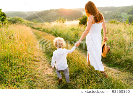 Happy mother with her son are walking at sunset along a field path holding hands. 92560590