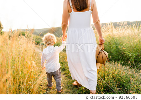 Woman in a beautiful white dress and her cute curly-haired son are walking in a summer field back view Woman in a beautiful white dress and her cute curly-haired son are walking in a summer field back view 92560593
