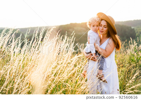 Portrait of a beautiful woman with her son in her arms in a field at sunset. 92560616