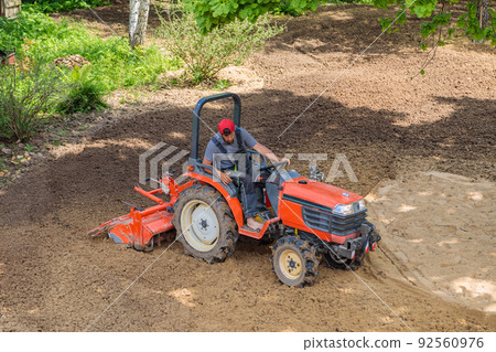 A farmer on a mini tractor loosens the soil for the lawn. Land cultivation, surface leveling A farmer on a mini tractor loosens the soil for the lawn. Land cultivation, surface leveling 92560976