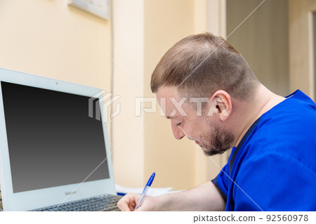 A male ophthalmologist sits at a workplace at a computer and writes with a pen 92560978
