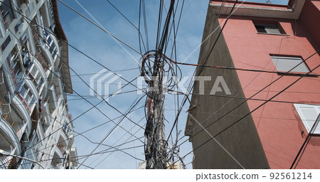 Tangled electricity fiber optic cables in communication systems, against blue sky, residential apartment buildings. Mess in black wires of power supply on electrical pole. Telecommunications problems Tangled electricity fiber optic cables in communication systems, against blue sky, residential apartment buildings. Mess in black wires of power supply on electrical pole. Telecommunications problems 92561214
