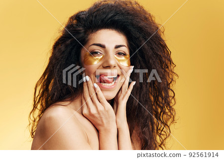 Enjoyed happy pretty Latin female with gold hydrogel patches under eye, stick tongue touch cheek posing isolated over yellow background. Cosmetic product ad Natural beauty concept Studio portrait Enjoyed happy pretty Latin female with gold hydrogel patches under eye, stick tongue touch cheek posing isolated over yellow background. Cosmetic product ad Natural beauty concept Studio portrait 92561914