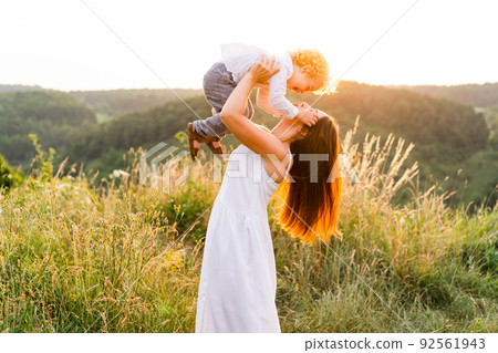 Playful mother with her son in a field at sunset. Happy woman throws boy up while walking in nature Playful mother with her son in a field at sunset. Happy woman throws boy up while walking in nature 92561943