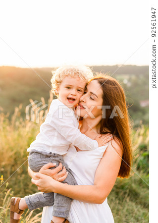 Happy mother hugs her little son, whom she holds in arms while walking in a summer field Happy mother hugs her little son, whom she holds in arms while walking in a summer field 92561947