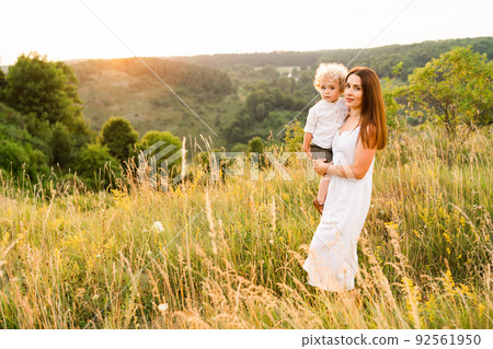 Portrait of a beautiful woman in a white dress with a cute curly boy in her arms in nature 92561950