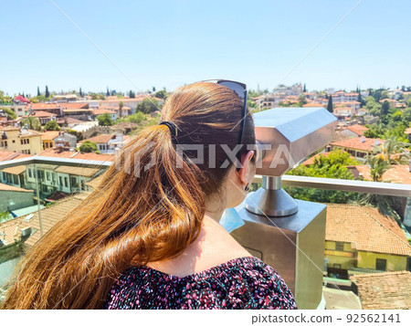 binoculars for tourists are installed on the roof. a girl with long, dark hair in a ponytail looks at houses with tiled roofs. woman on a sightseeing tour in a tropical country binoculars for tourists are installed on the roof. a girl with long, dark hair in a ponytail looks at houses with tiled roofs. woman on a sightseeing tour in a tropical country 92562141
