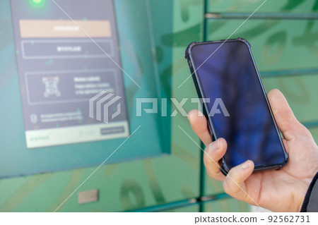 Female hand holding mobile phone next to Parcel locker Collecting parcel from shopping locker. Woman Skans QR Code on Mobile phone Self-service Locker Cell with Bar code Reader Modern Shipping and 92562731