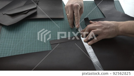 Close up of leather craftsman working with natural leather by knife and ruler by cutting a leather to a peaces. Handmade master at work in local workshop. Leather wallet stitch. 92562825