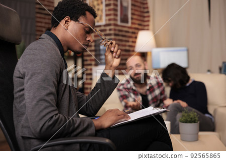 Stressed counselor taking notes on paper at couple therapy session with married partners, having relationship conflict and difficulties. Trying to solve problems with psychology advice. Stressed counselor taking notes on paper at couple therapy session with married partners, having relationship conflict and difficulties. Trying to solve problems with psychology advice. 92565865