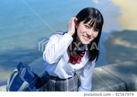 一個坐在美麗的海邊防波堤上的女高中生，梳著頭髮，眼中帶著笑意。放學後，在海灘 92567979