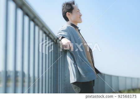 A man relaxing on a fence in a park by the pond. 92568175