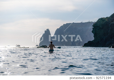 Side view foto of a man swiming and relaxing on the sup board. Sportive man in the sea on the Stand Up Paddle Board SUP. The concept of an active and healthy life in harmony with nature. 92571194