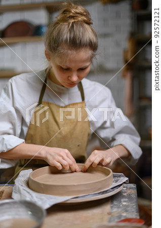 A ceramist makes a plate. Woman in an apron works in a pottery workshop. A ceramist makes a plate. Woman in an apron works in a pottery workshop. 92572121