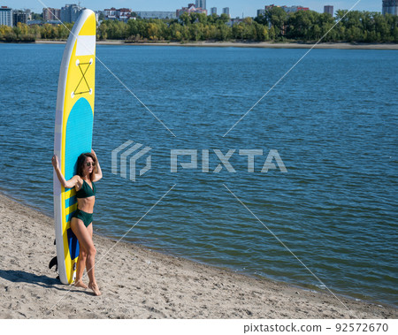 Caucasian woman walks along the beach and carries a sup board on the river in the city. Summer sport. 92572670