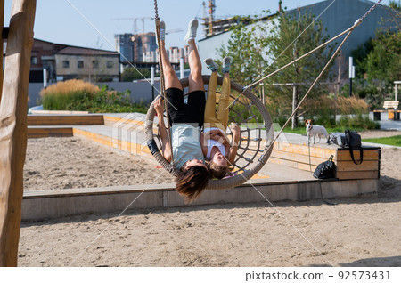 Mom and daughter swing on a round swing. Caucasian woman and little girl have fun on the playground. 92573431