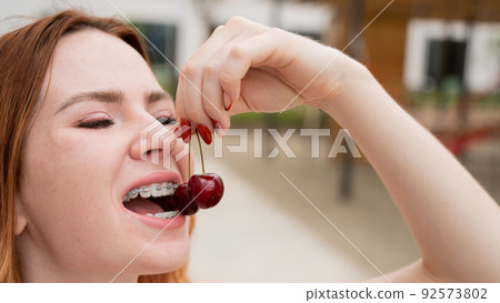 Beautiful young red-haired woman with braces on her teeth eats sweet cherries in the summer outdoors Beautiful young red-haired woman with braces on her teeth eats sweet cherries in the summer outdoors 92573802