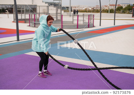 Caucasian woman in a mint sweatshirt is training with battle ropes at the sports ground.  92573876