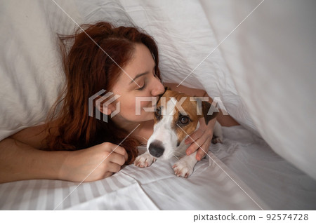 Caucasian red-haired woman sleeps in an embrace with a jack russell terrier dog on a white sheet. 92574728