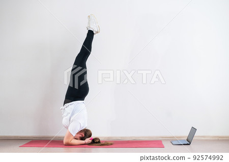 A chubby young woman watches an online yoga lesson on a laptop. Distance sports training. Forearm stand A chubby young woman watches an online yoga lesson on a laptop. Distance sports training. Forearm stand 92574992