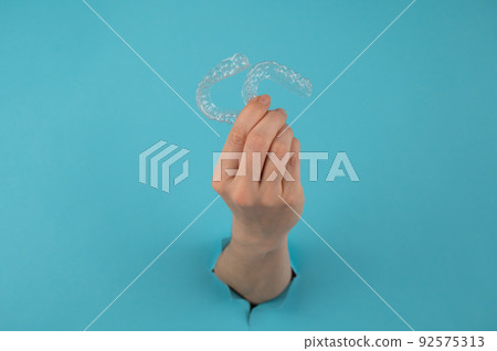 A female hand sticking out of a hole from a blue background holds removable night retainers.  92575313