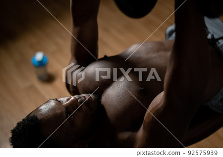 Shirtless afro american man doing exercises with dumbbells lying on bench in gym.  92575939