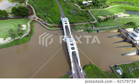 Aerial view of Tha Chomphu White Bridge, Lamphun, Thailand with river, forest trees and green mountain hill. An old railway bridge across the river. Tourist attraction landmark. 92579739