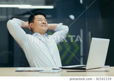 A break at work. A young Asian man is resting at the workplace at the desk in the office. He sits with his hands behind his head, closes his eyes, rests. 92581159